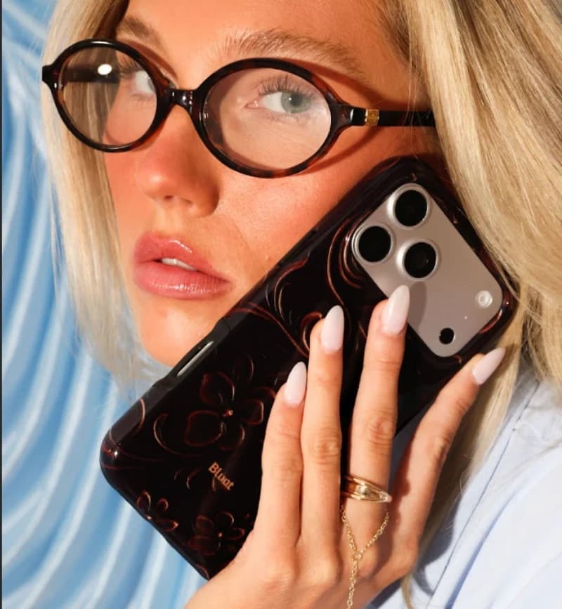 A blonde girl in tortoiseshell glasses holding a tortoiseshell-print Blunt case with floral swirls to her cheek
