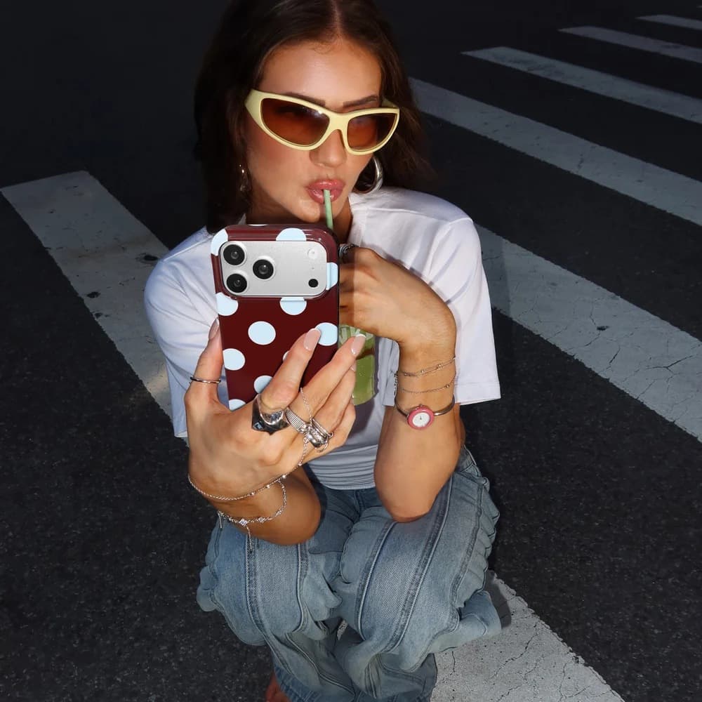 A girl in yellow wraparound sunglasses taking a mirror selfie with a burgundy and baby-blue polka dot Velvet Caviar case