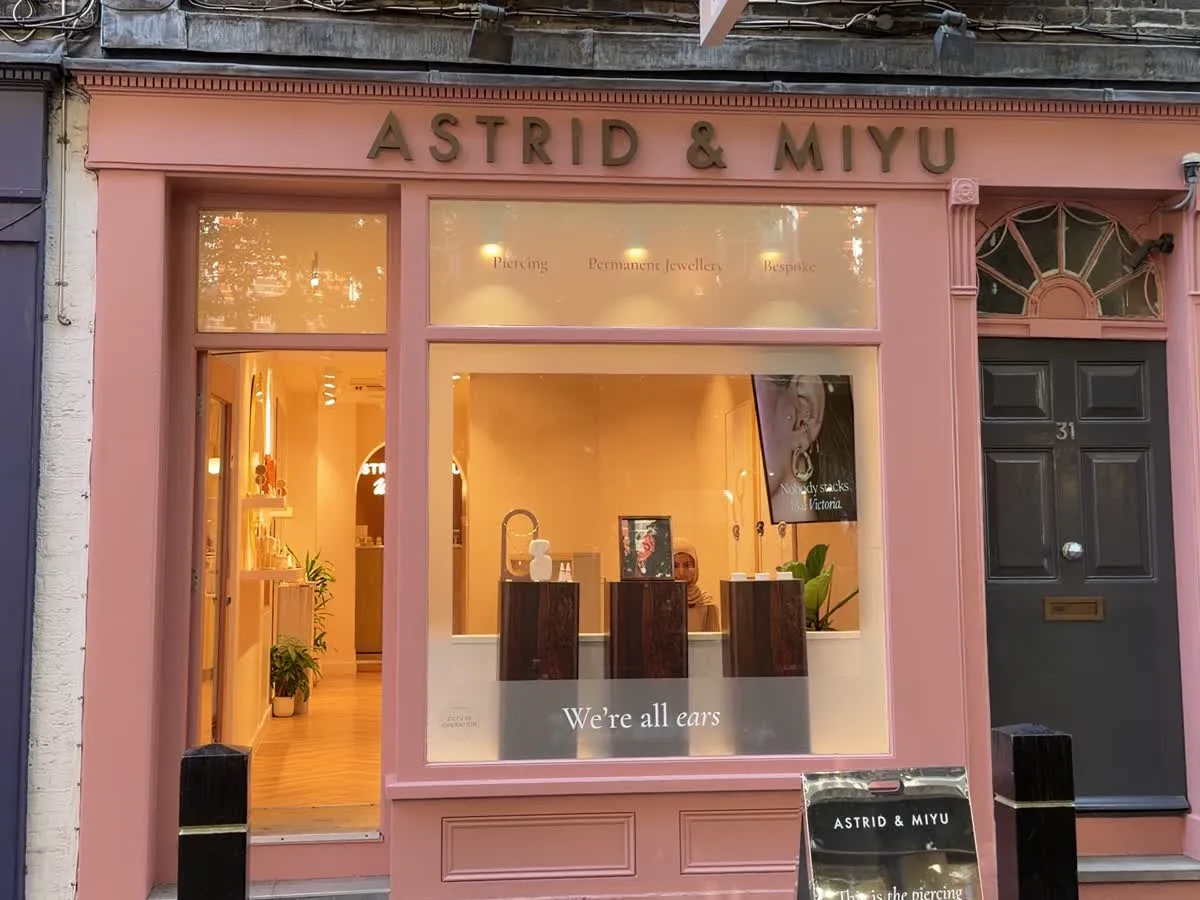 The Astrid & Miyu shop: a dusty-pink Georgian shopfront with the name in gold, a warmly lit window reading “we're all ears”, and a pavement board advertising piercing